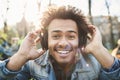 Portrait of positive adult dark-skinned man smiling broadly while sitting in park, listening to music in headphones and