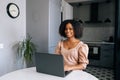 Portrait of positive African young woman sitting at table in modern kitchen room working on laptop, smiling looking at Royalty Free Stock Photo