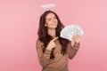 Portrait of positive angelic young woman with halo above head pointing at dollar banknotes, showing big money