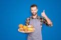 Portrait of positive attractive baker in uniform with stubble holding tray with bakery products in hand, showing thumb Royalty Free Stock Photo