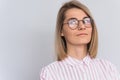 Portrait of positive blonde female with attractive look wearing pink shirt and round trendy glasses, posing against white wall in