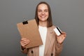 Portrait of positive cheerful woman with brown hair wearing beige jacket, standing with documents and showing credit card, looking Royalty Free Stock Photo