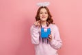 Portrait of positive curly haired teenage girl in hoodie with halo on head holding wrapped gift box and pointing finger on camera Royalty Free Stock Photo