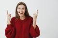 Portrait of positive joyful charming woman in red loose sweater, raising index fingers and pointing up, looking aside