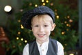 Portrait of positive little boy in cap against backdrop of Christmas tree with garland lights. Sincere child emotions. Happy child