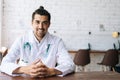 Portrait of positive male doctor wearing white uniform with stethoscope sitting at desk in hospital office, smiling Royalty Free Stock Photo