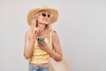 Portrait positive mature blond woman in straw hat, yellow top and sunglasses holding smartphone and shopping bag isolated on white
