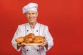 Portrait of positive senior attractive baker in uniform having tray with bakery products, isolated on red background