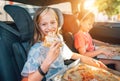 Portrait of positive smiling girl eating just cooked italian pizza sitting with a sister on car back seat in child car seats.