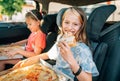 Portrait of positive smiling girl eating just cooked italian pizza sitting with a sister on car back seat in child car seats. Royalty Free Stock Photo