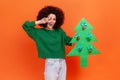 Portrait of positive woman with Afro hairstyle wearing green casual style sweater holding paper christmas tree and showing v sign Royalty Free Stock Photo