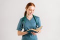 Portrait of positive young woman physician in green uniform with stethoscope reading medical book standing on white