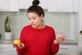 Portrait of pragnant woman deciding to choose healthy or unhealthy food, posing  on white kitchen interior background, Royalty Free Stock Photo