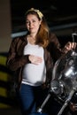 Portrait of pregnant woman dressed in brown leather jacket standing in front of motorcycle headlight and holding large belly Royalty Free Stock Photo