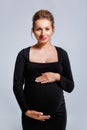 Portrait of a pregnant woman in a tight black dress on a gray background. The woman gently holds her stomach with her hands Royalty Free Stock Photo