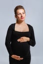 Portrait of a pregnant woman in a tight black dress on a gray background. The woman gently holds her stomach with her hands Royalty Free Stock Photo