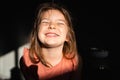 Portrait of preschool age girl in sunlight and dark background. Happy smiling emotions and closed eyes