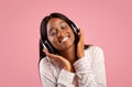 Portrait of pretty African American woman wearing headphones and listening to beautiful music on pink studio background Royalty Free Stock Photo