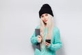 Portrait of pretty blonde girl with laptop in hands using smartphone on white background. Wearing black hat and blue sweater. Royalty Free Stock Photo