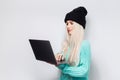 Portrait of pretty blonde girl using laptop on white background. Wearing blue sweater and black hat. Royalty Free Stock Photo