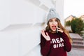 Portrait pretty brunette with long hair in knitted hat and marsala sweater near grey wall on street.