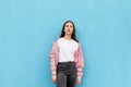 Portrait of pretty caucasian teen girl in plain white t-shirt and pastel pink bomber jacket standing against blue city