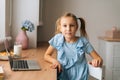 Portrait of pretty elementary child girl sitting at home table with laptop and snack by window, looking at camera.
