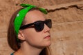 Portrait of pretty hipster girl in sunglasses, green sweatshirt, bandana standing on clay rock background on sunny day
