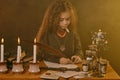 Little witch dressed in dark clothes sitting at the table against a black smoky background. There are magic wand, books Royalty Free Stock Photo