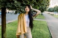 Portrait of pretty lovely Asian girl with long dark hair, posing on street on background of city park alley with green