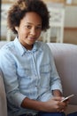 Portrait of pretty mixed race teen girl in earphones looking at camera while sitting on the couch and using mobile phone Royalty Free Stock Photo