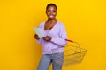 Portrait of pretty positive girl hold shopping list empty basket isolated on yellow color background