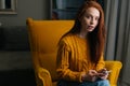 Portrait of pretty red-haired young woman using smartphone sitting in yellow armchair, confident looking at camera. Royalty Free Stock Photo