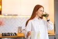 Portrait of pretty redhead young woman drinking healthy vegetable detox smoothie juice from blender standing in kitchen Royalty Free Stock Photo