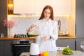 Portrait of pretty redhead young woman pouring green vegetable detox smoothie cocktail from blender into glass in Royalty Free Stock Photo