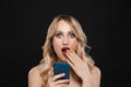 Shocked young blonde woman with bright makeup red lips posing isolated over black wall background using mobile phone Royalty Free Stock Photo