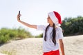 Portrait of a pretty smiling teenage girl in a red Santa hat and taking selfie on a sand tropical beach Royalty Free Stock Photo