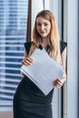 Portrait of pretty smiling young woman holding papers standing at window with cityscape view Royalty Free Stock Photo