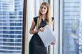 Portrait of pretty smiling young woman holding papers standing at window with cityscape view Royalty Free Stock Photo