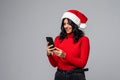 Portrait of a pretty woman dressed in red christmas hat holding mobile phone while standing and looking at camera isolated over Royalty Free Stock Photo