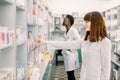 Portrait of a pretty woman pharmacist searching medicines on the shelf at the drugstore, African male colleague working