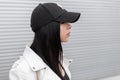 Portrait of pretty young beautiful woman in a stylish white leather jacket in a fashionable black baseball cap near a metal wall Royalty Free Stock Photo