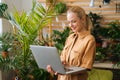 Portrait of pretty young businesswoman floral store owner standing with laptop holding in hand among green plants