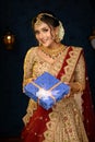 Portrait of a pretty young Indian woman dressed in traditional lehenga, gold jewellery and bangles holding gift box in hands on Royalty Free Stock Photo