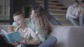 Portrait of pretty young mother reading book with her son sitting on the sofa while her two teen kids sitting on the