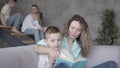 Portrait of pretty young mother reading book to her son while the rest of her teen kids playing with each other in the