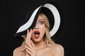 Shocked blonde woman with bright makeup red lips posing isolated over black wall background wearing hat
