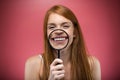 Pretty young woman showing teeth trought magnifying glass over pink background.