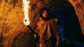 Portrait of Primeval Caveman Wearing Animal Skin Exploring Cave At Night, Holding Torch with Fire