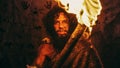 Portrait of Primeval Caveman Wearing Animal Skin Standing in His Cave At Night, Holding Torch with